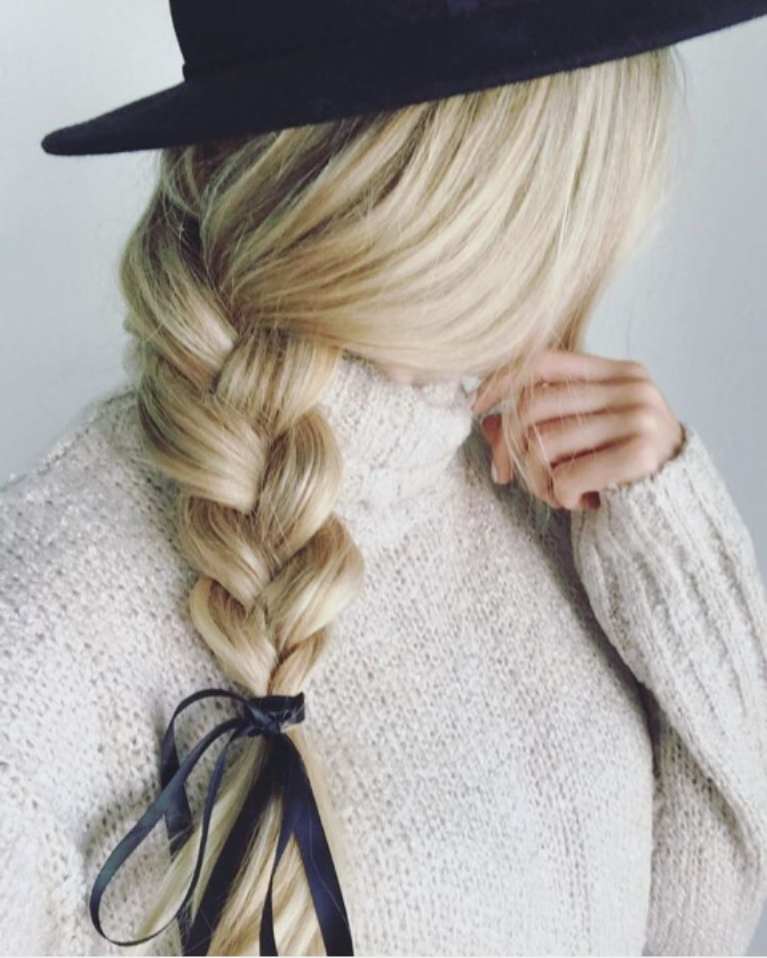 A thick blonde braid tied with a black ribbon, worn with a light sweater and dark hat.